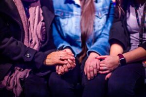 Three women holding hands, symbolizing support and empathy in a warm setting.