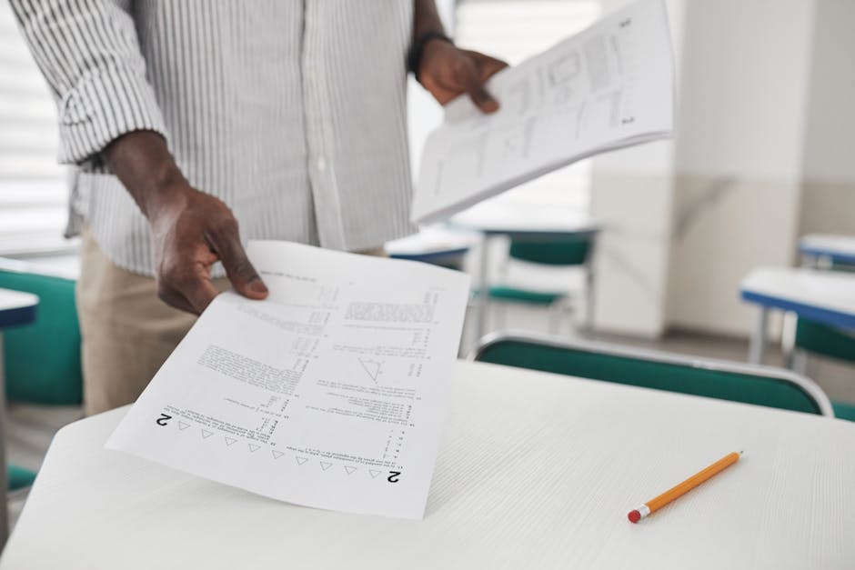 Close-up of a person arranging examination papers on a desk in a classroom.