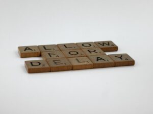 Wooden Scrabble tiles arranged on a white surface spelling 'Allow for Delay'.