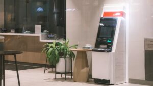 An ATM machine stands in a modern bank lobby, next to a plant and table.