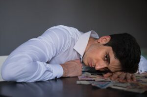 Adult man in shirt lying on table, surrounded by currency, looking stressed indoors.